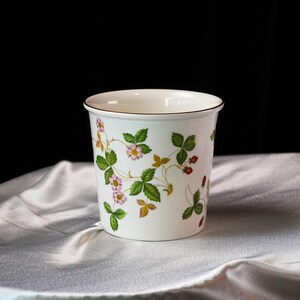 Op de afbeelding: Witte keramische plantenpot met een bruine rand, versierd met een aardbeienplantmotief. Het ontwerp bevat groene bladeren, roze bloemen en rode aardbeien. De pot staat op een witte, getextureerde stof.