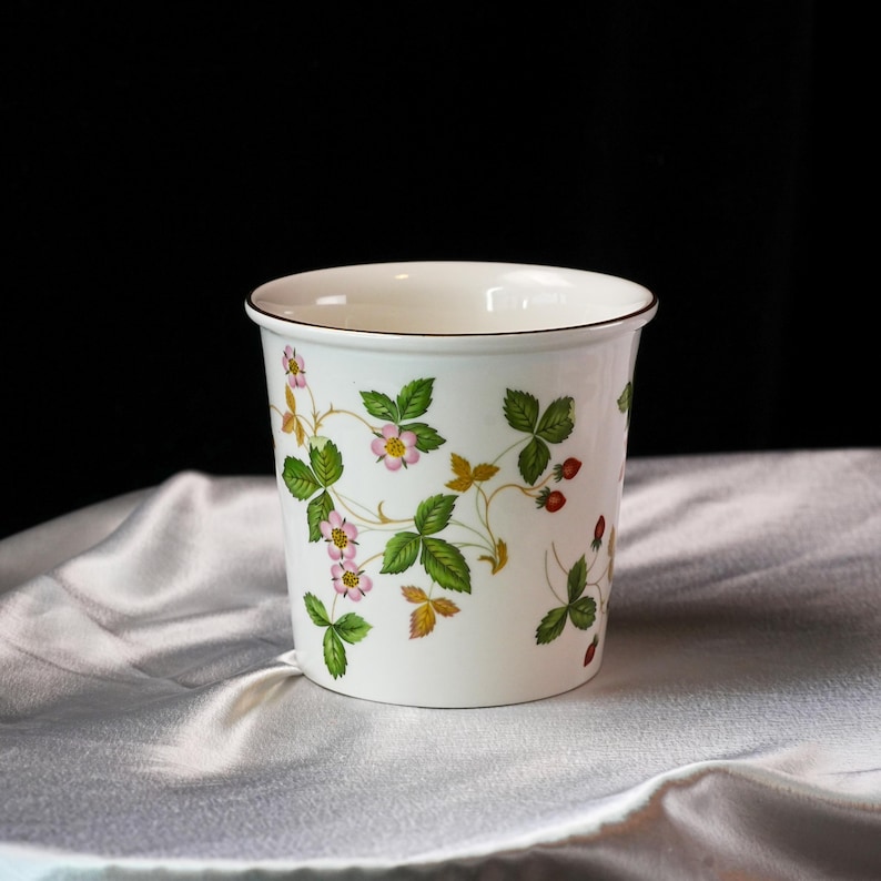 Op de afbeelding: Witte keramische plantenpot met een bruine rand, versierd met een aardbeienplantmotief. Het ontwerp bevat groene bladeren, roze bloemen en rode aardbeien. De pot staat op een witte, getextureerde stof.