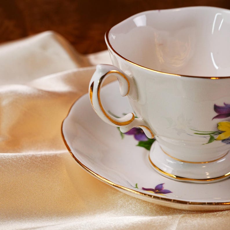 K&ouml;nnte beinhalten: Ein wei&szlig;es Tee-Set mit goldfarbenen R&auml;ndern und Blumendekor. Die Tasse hat einen geschwungenen Henkel und steht auf einer passenden Untertasse. Das Set befindet sich auf einem cremefarbenen Stoff.