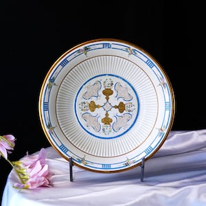 Può includere: Un piatto bianco decorativo con bordo dorato e dettagli blu. Il piatto presenta un disegno centrale con motivi floreali dorati e blu. Il piatto è esposto su un supporto metallico contro un tessuto bianco.