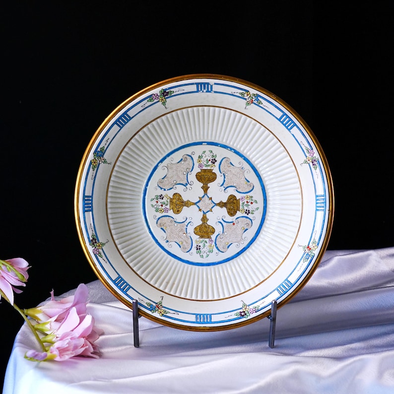 May include: A decorative white plate with a gold rim and blue accents. The plate features a central design with gold and blue floral patterns. The plate is displayed on a metal stand against a white cloth.