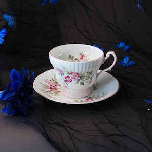 May include: A white porcelain teacup and saucer set featuring a delicate ribbed texture and a floral pattern of pink wild roses with green leaves. The rim of the cup and the edge of the saucer are accented with a thin, soft pink painted line.