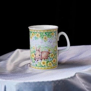 Puede incluir: Una taza de cer&aacute;mica blanca con una colorida ilustraci&oacute;n de un conejo marr&oacute;n entre flores de primavera amarillas y campanillas de color p&uacute;rpura claro. El dise&ntilde;o incluye un borde decorativo amarillo y verde en los bordes superior e inferior.