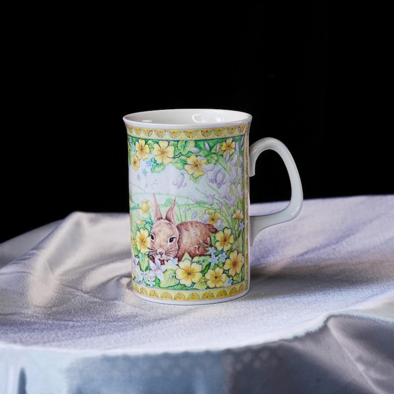 Puede incluir: Una taza de cer&aacute;mica blanca con una colorida ilustraci&oacute;n de un conejo marr&oacute;n entre flores de primavera amarillas y campanillas de color p&uacute;rpura claro. El dise&ntilde;o incluye un borde decorativo amarillo y verde en los bordes superior e inferior.
