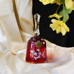 May include: A red glass bell with a clear handle and gold detailing. The bell is decorated with hand-painted blue and white floral patterns and a gold rim. A gold label is attached to the bell.