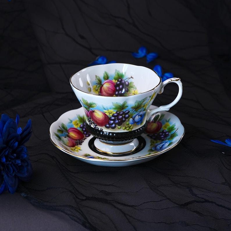 Op de afbeelding: Een witte porseleinen theekop en schotel met een kleurrijk fruitpatroon van druiven, perziken en gele bloemen. De voet van de kop en het midden van de schotel zijn zwart met een geribbelde textuur en afgewerkt met een gouden randje.
