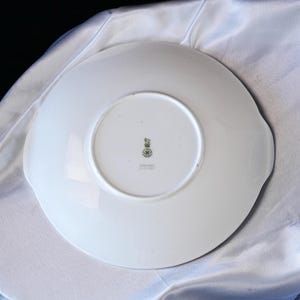 May include: White ceramic plate with a scalloped edge and two small handles. The underside features a green and gold emblem and text. The plate is set against a white, textured background.