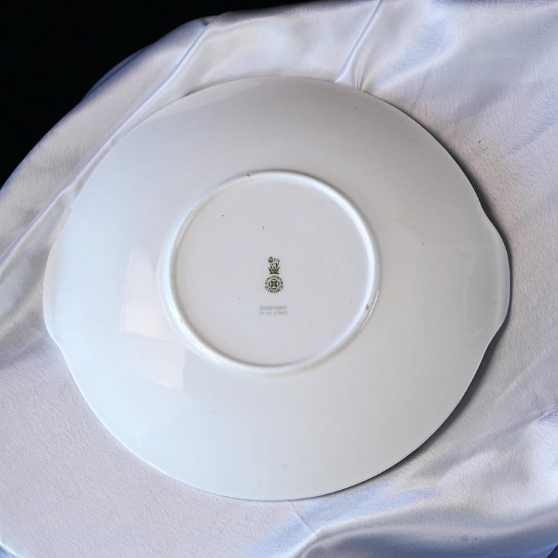 May include: White ceramic plate with a scalloped edge and two small handles. The underside features a green and gold emblem and text. The plate is set against a white, textured background.