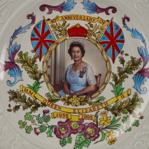 Op de afbeelding: Een wit keramieken herdenkingsbord met een centraal portret van Koningin Elizabeth II. De tekst luidt 40th ANNIVERSARY OF HM QUEEN ELIZABETH II 1952 1992. Het ontwerp bevat twee Union Jack vlaggen, een rode kroon en kleurrijke bloemmotieven.