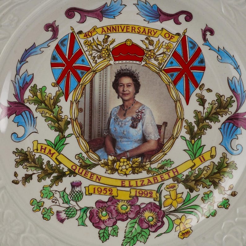 Op de afbeelding: Een wit keramieken herdenkingsbord met een centraal portret van Koningin Elizabeth II. De tekst luidt 40th ANNIVERSARY OF HM QUEEN ELIZABETH II 1952 1992. Het ontwerp bevat twee Union Jack vlaggen, een rode kroon en kleurrijke bloemmotieven.