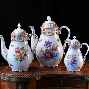 May include: A set of three white porcelain teapots and a creamer, each featuring colorful hand-painted floral bouquets, gold-trimmed rims, and matching lids with finials. The vessels have curved handles and spouts, showcasing intricate botanical patterns in shades of pink, orange, blue, and green against a glossy white finish.