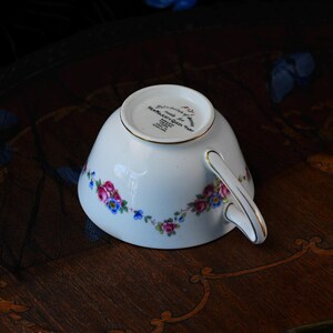 Op de afbeelding: Een witte porseleinen theekop met een goudkleurig handvat en een bloemmotief van roze rozen en blauwe bloemen. De onderkant bevat zwarte tekst: Made for Her Majesty Queen Mary, Paragon Fine Bone China, Made in England en F121.