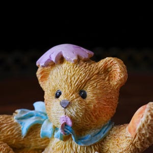 May include: A detailed figurine of a teddy bear with a light brown fur texture. The bear wears a light blue ribbon around its neck and a purple flower hat. The bear is holding a small pink flower in its mouth.