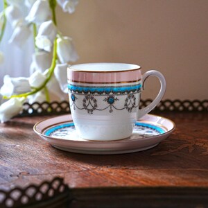 May include: A white teacup and saucer set with a pink rim and gold trim. The cup and saucer feature a blue and black decorative band. The set is placed on a wooden tray.