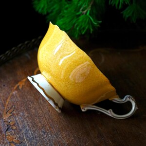 May include: A yellow teacup with a white base and a silver handle. The teacup has a scalloped rim and a gold trim. The teacup is resting on a wooden surface.