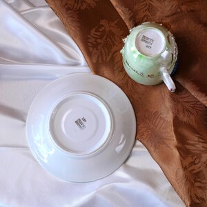 May include: A white porcelain teacup and saucer set. The teacup is decorated with green, blue, and gold accents. The saucer is plain white. The set is displayed on a brown and white fabric background. The bottom of the saucer and teacup have text.