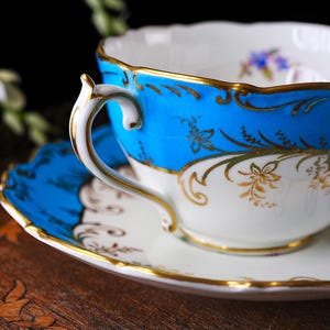Op de afbeelding: Een vintage theekop en schotel set. De theekop heeft een helderblauwe rand met gouden bloemmotieven en een witte binnenkant. De schotel weerspiegelt het blauwe en gouden ontwerp. Het handvat is wit met gouden rand.