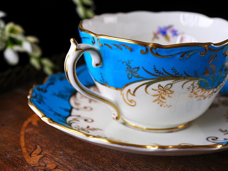 Op de afbeelding: Een vintage theekop en schotel set. De theekop heeft een helderblauwe rand met gouden bloemmotieven en een witte binnenkant. De schotel weerspiegelt het blauwe en gouden ontwerp. Het handvat is wit met gouden rand.