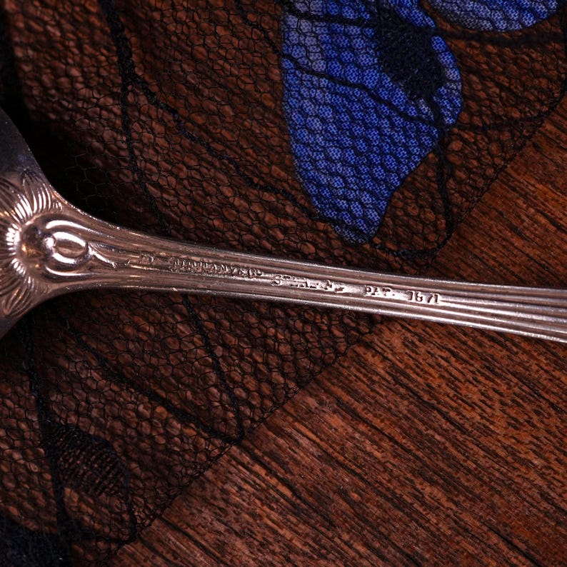 Puede incluir: Un mango de cuchara de metal plateado con un dise&ntilde;o floral en relieve en el cuello. El mango est&aacute; grabado con el texto Tiffany & Co. y Pat. 1871. La superficie met&aacute;lica muestra un acabado reflectante con estr&iacute;as longitudinales visibles.