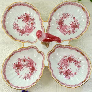 May include: A white porcelain serving dish with four shell-shaped sections, each featuring a pink floral pattern and gold edging. A red and white bird figurine is centrally placed, enhancing the decorative appeal of this elegant serving piece.