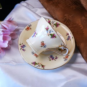 Op de afbeelding: Cr&egrave;mekleurige theekop en schotel set met bloemmotieven in roze, blauw en oranje. De kop is gekanteld op de schotel, wat het delicate ontwerp en de gouden rand benadrukt. Een klassieke vintage theeset.