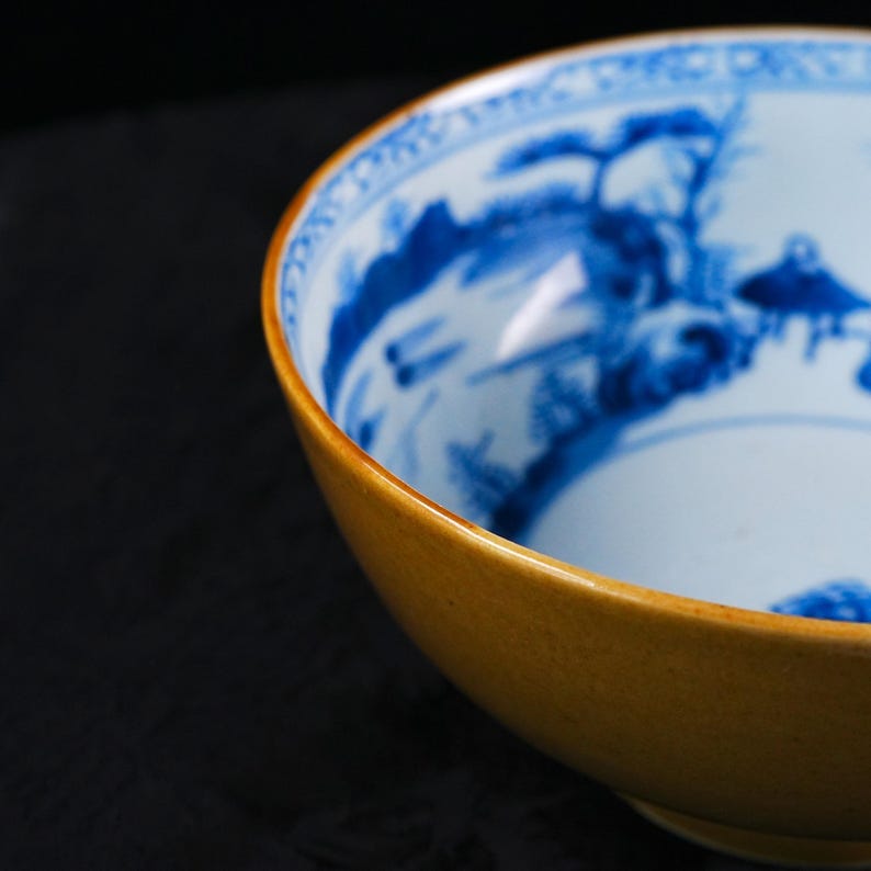 May include: A ceramic bowl with a blue and white landscape design on the inside. The exterior of the bowl is a warm, golden-yellow color. The bowl's rim has a subtle, decorative pattern. The bowl is likely for serving food or as a decorative piece.