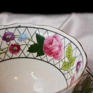 Op de afbeelding: Close-up van een witte theekop met een bloemenontwerp. De rand heeft een zwart geometrisch patroon met roze rozen, paarse en blauwe bloemen en groene bladeren. De kop heeft een gouden rand.