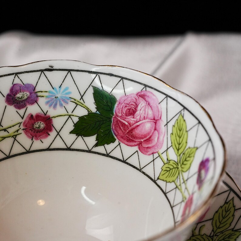Op de afbeelding: Close-up van een witte theekop met een bloemenontwerp. De rand heeft een zwart geometrisch patroon met roze rozen, paarse en blauwe bloemen en groene bladeren. De kop heeft een gouden rand.