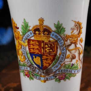 Puede incluir: Una taza de cer&aacute;mica blanca con un escudo real colorido que incluye una corona, un le&oacute;n y un unicornio. El dise&ntilde;o presenta el texto Coronation A.D. 1911 George V. R. y una liga azul con la inscripci&oacute;n Honi Soit Qui Mal Y Pense.
