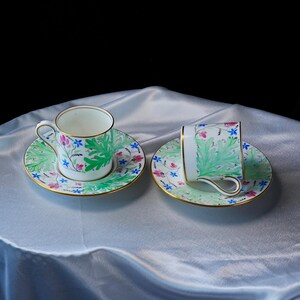 May include: Two white porcelain teacups and matching saucers featuring a hand-painted botanical pattern of large green leaves, small blue flowers, and pink buds. Each piece is finished with a thin gold rim and a curved handle on the teacups.