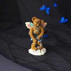 Puede incluir: Una figura de oso de peluche marr&oacute;n con pelaje texturizado, que lleva una tela azul claro alrededor de la cintura y alas multicolores. El oso se apoya sobre una base blanca en forma de nube con una peque&ntilde;a flecha plateada y un carcaj.