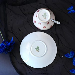 Może przedstawiać: Zestaw białej porcelanowej filiżanki ze spodkiem, ozdobiony delikatnym wzorem r&oacute;żowych r&oacute;ż. Sp&oacute;d obu element&oacute;w posiada zieloną sygnaturę Aynsley England z napisem C1312/3. Filiżanka ma srebrzysty uchwyt oraz wykończenie krawędzi.
