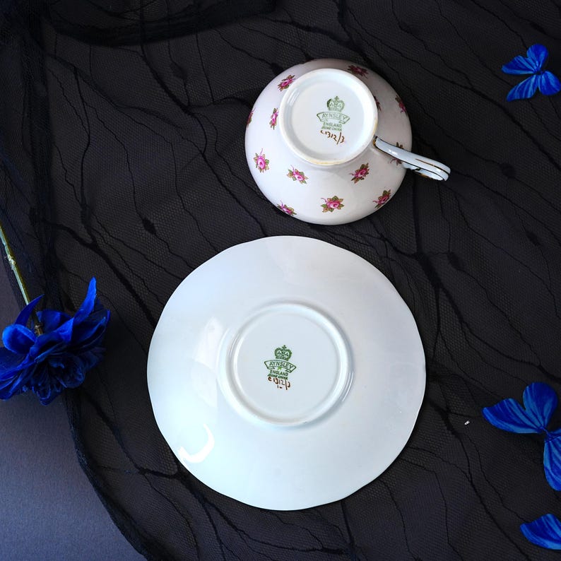 Może przedstawiać: Zestaw białej porcelanowej filiżanki ze spodkiem, ozdobiony delikatnym wzorem r&oacute;żowych r&oacute;ż. Sp&oacute;d obu element&oacute;w posiada zieloną sygnaturę Aynsley England z napisem C1312/3. Filiżanka ma srebrzysty uchwyt oraz wykończenie krawędzi.