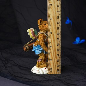 Puede incluir: Una figura de oso de peluche texturizado marr&oacute;n con alas multicolores, una tela azul y un carcaj de madera, sobre una base de nube blanca. Una regla de madera junto a la figura muestra una altura de aproximadamente 11,5 cent&iacute;metros.