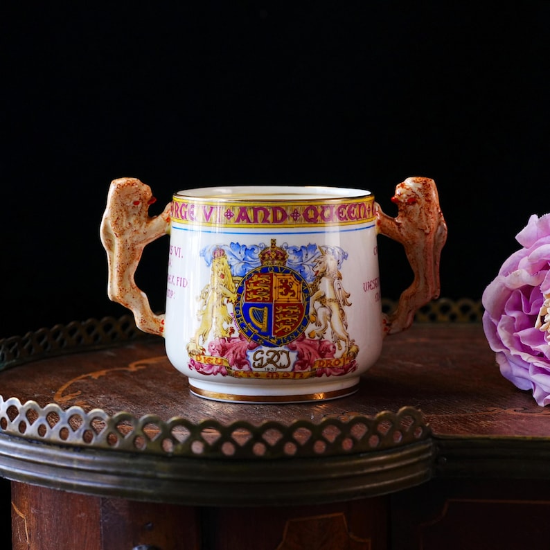Puede incluir: Una taza de cer&aacute;mica blanca con asas ornamentadas y un dise&ntilde;o de escudo real. La taza presenta un borde dorado y el texto "GEORGE VI AND QUEEN ELIZABETH". Las asas est&aacute;n esculpidas en un estilo texturizado &uacute;nico. Una flor rosa es visible en el fondo.