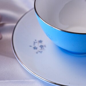 Pode incluir: Conjunto de ch&aacute; azul claro com acabamento prateado. A ch&aacute;vena est&aacute; cheia de branco e assenta num pires a condizer. O pires tem um delicado desenho floral em azul.