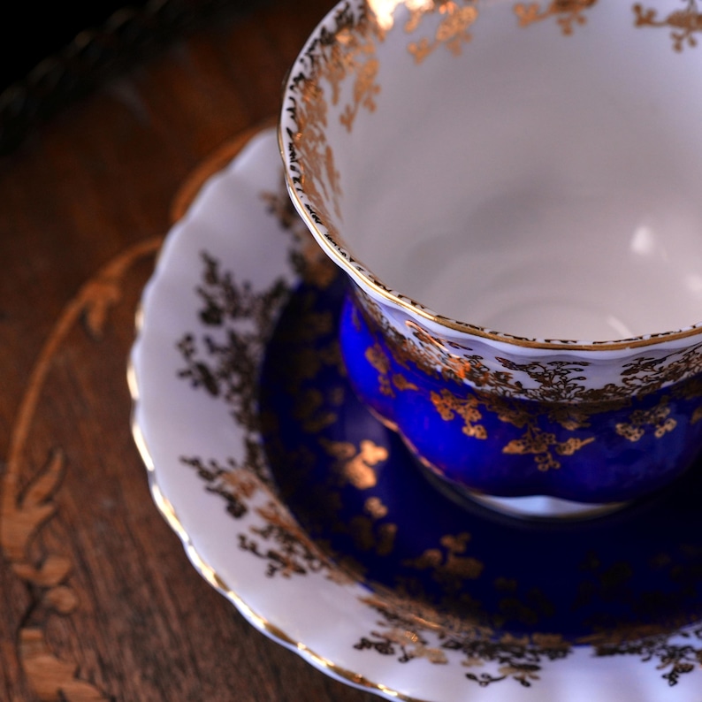 May include: A detailed shot of a vintage teacup and saucer. The teacup features a white interior and a deep blue exterior, adorned with gold floral patterns. The saucer, with a white rim and gold accents, complements the blue centre. Set on a wooden surface.
