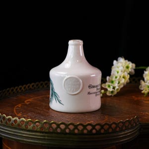 K&ouml;nnte beinhalten: Eine wei&szlig;e Keramikflasche mit rundem K&ouml;rper und schmalem Hals. Die Flasche hat ein rundes Emblem und Text auf der Seite sowie ein gr&uuml;nes botanisches Design. Sie steht auf einer Holzoberfl&auml;che mit dekorativem Metallrand.