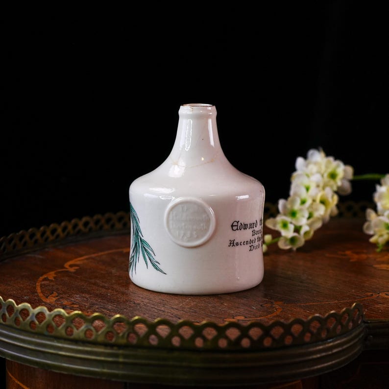 K&ouml;nnte beinhalten: Eine wei&szlig;e Keramikflasche mit rundem K&ouml;rper und schmalem Hals. Die Flasche hat ein rundes Emblem und Text auf der Seite sowie ein gr&uuml;nes botanisches Design. Sie steht auf einer Holzoberfl&auml;che mit dekorativem Metallrand.