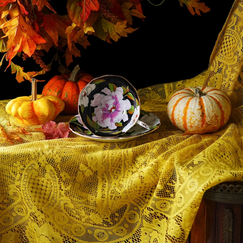 May include: A black teacup and saucer set with a floral design featuring a large white and pink peony. The teacup and saucer are set on a yellow lace tablecloth, with pumpkins and autumn leaves in the background.