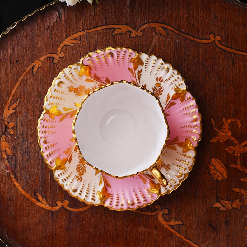 May include: A vintage teacup and saucer set. The teacup is white with a gold handle. The saucer is pink and white with gold floral accents. The set is placed on a dark wooden surface with decorative trim.