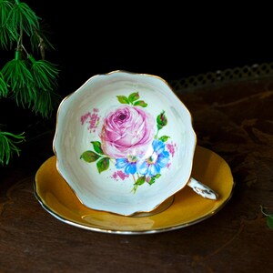 May include: A vintage teacup and saucer set. The teacup features a pink rose and blue flower design with gold trim on a white background. The saucer is a solid yellow colour with a gold rim. The set is placed on a dark wooden surface.