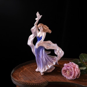 May include: A porcelain figurine of a woman in a flowing blue and purple gown, holding a white dove. Her auburn hair cascades down her shoulders, and a sheer scarf drapes around her. The figurine is set on a wooden table with a pink rose.