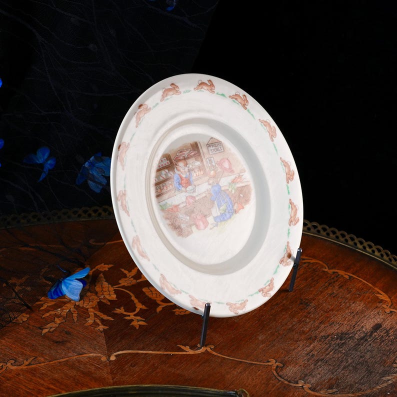 May include: A round ceramic plate featuring a central illustration of two rabbits in a shop setting with a sign reading 'P. RABBIT'S STORE'. The rim is decorated with a repeating pattern of small brown rabbits and green grass accents.