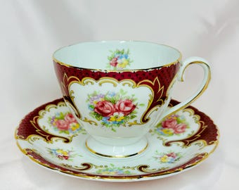 May include: A white teacup and saucer with a burgundy floral pattern and gold accents. The cup and saucer feature a delicate floral design with pink and yellow roses, and a gold trim around the edges.