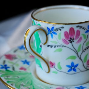 May include: A white porcelain teacup and saucer set featuring hand-painted floral motifs in pink, blue, and green. The cup has a curved handle with gold trim along the rim and handle edges. The saucer displays matching floral patterns.