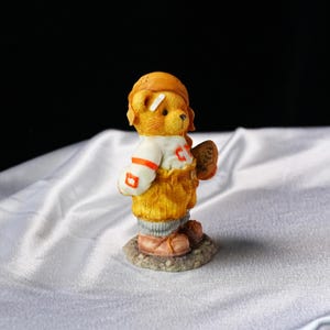 Op de afbeelding: Een gedetailleerd beeldje van een teddybeer in een honkbaluniform. De beer draagt een helm, een trui met de letter "C" en houdt een honkbalhandschoen vast. Het beeldje staat op een kleine, getextureerde basis.