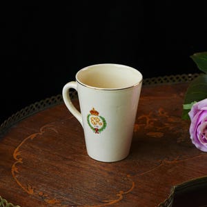 Puede incluir: Taza de cer&aacute;mica color crema con borde y asa dorados. La taza presenta un dise&ntilde;o de escudo con una corona, una corona de laurel y las iniciales "GR VIII" en rojo y dorado. La taza est&aacute; sobre una superficie de madera.