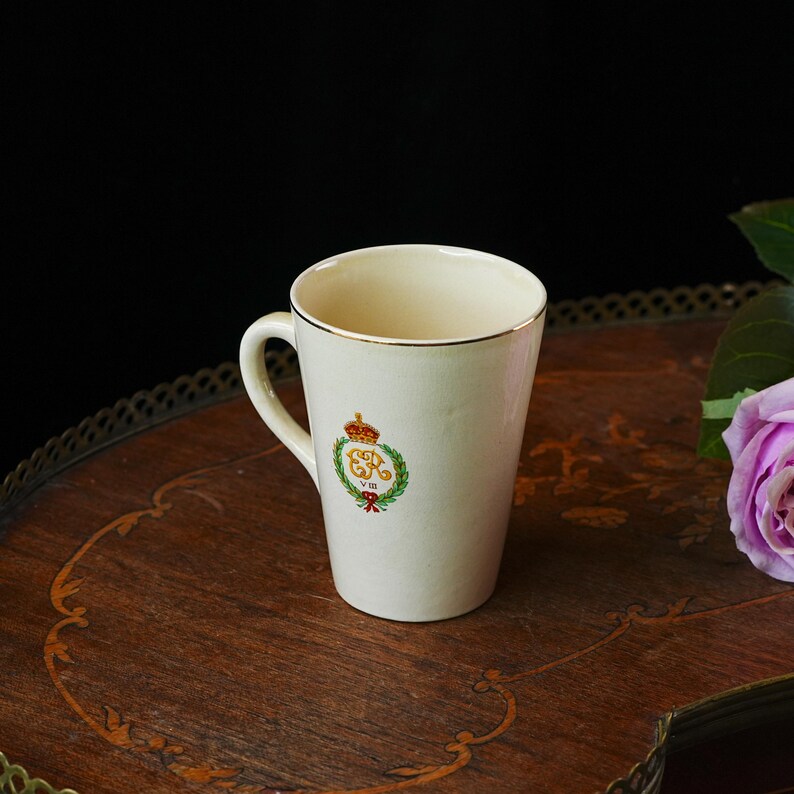 Puede incluir: Taza de cer&aacute;mica color crema con borde y asa dorados. La taza presenta un dise&ntilde;o de escudo con una corona, una corona de laurel y las iniciales "GR VIII" en rojo y dorado. La taza est&aacute; sobre una superficie de madera.