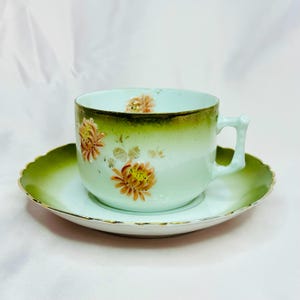 May include: A vintage teacup and saucer set with a green and white floral design. The cup has a delicate handle and the saucer has a scalloped edge.